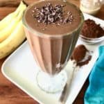 A vegan chocolate smoothie in a tall glass, topped with cacao nibs, on a white platter with bananas, cocoa powder and a jar of tahini