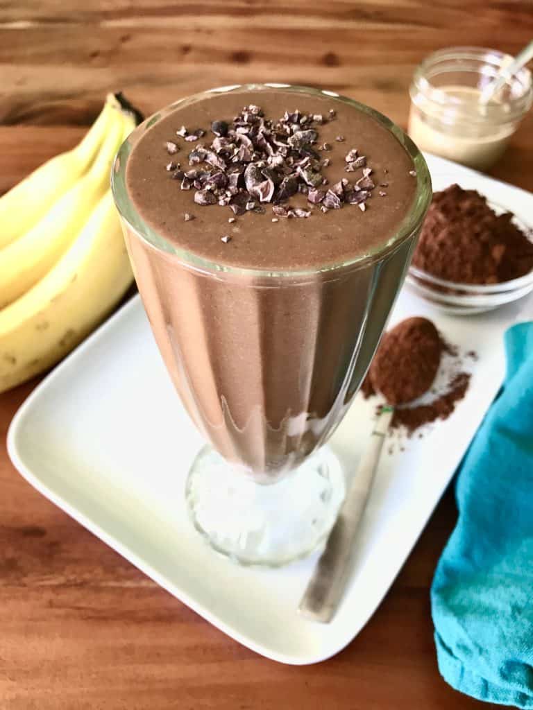 A vegan chocolate smoothie in a tall glass, topped with cacao nibs, on a white platter with bananas, cocoa powder and a jar of tahini