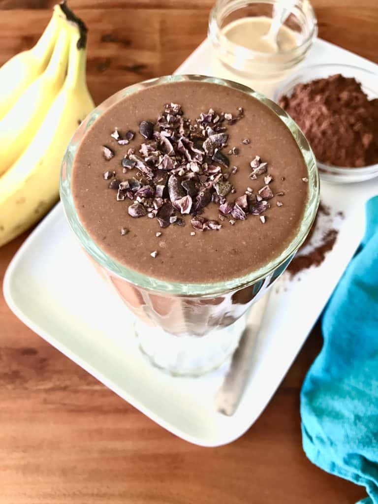 Chocolate Tahini Smoothie in a tall glass, topped with cacao nibs, on a white platter with bananas, cocoa powder and a jar of tahini