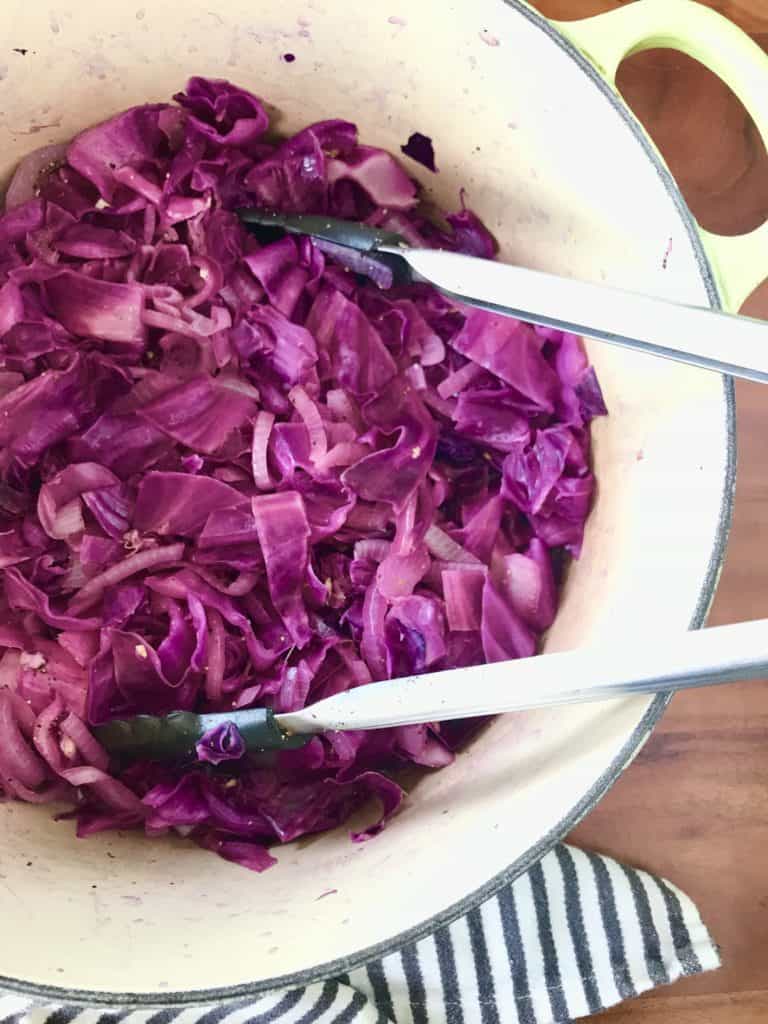 easy braised red cabbage in a big pot with tongs, next to a striped towel on a wooden table