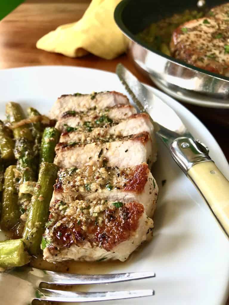one pan creamy mustard pork chops with asparagus on a white plate with the pan of chops and veggies behind it, all on a wooden table