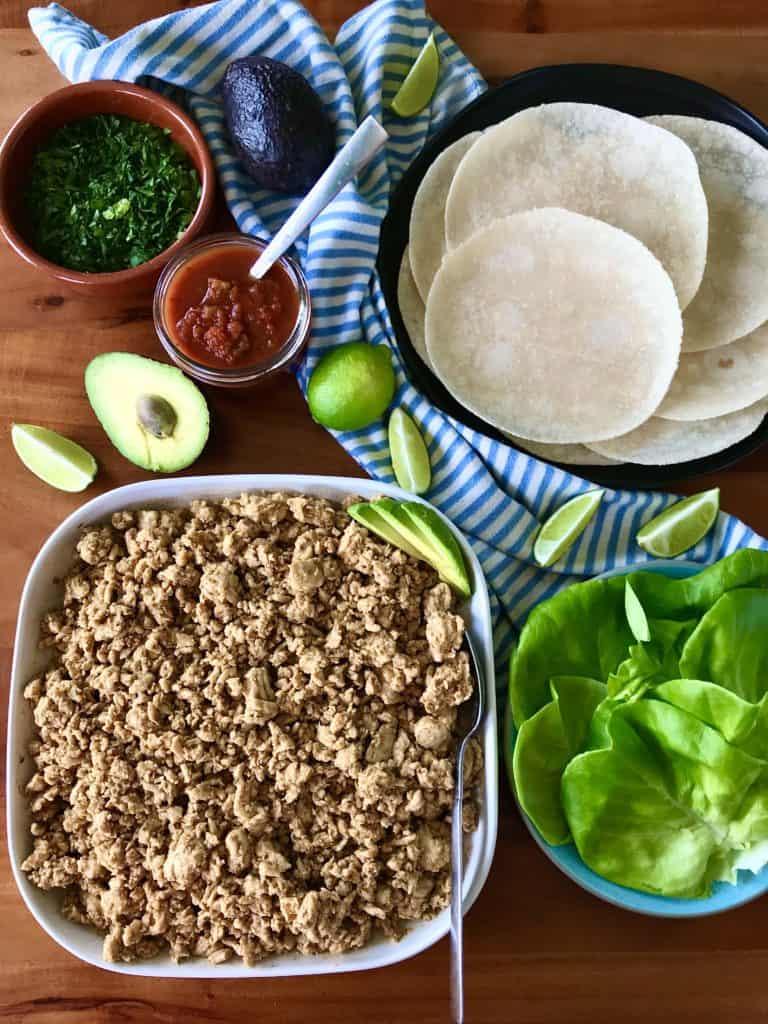 a wooden table with ground chicken taco meat, tortillas, lettuce, salsa, limes, avocado and a striped towel