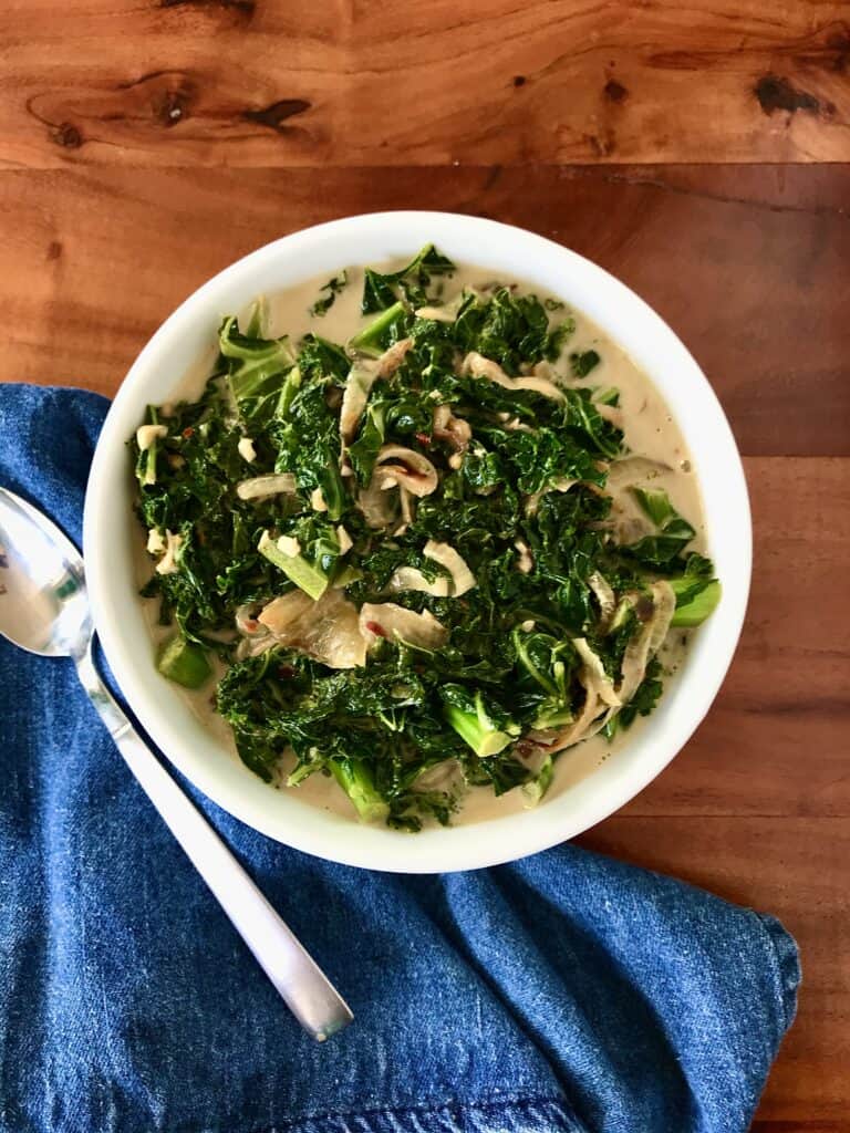 Whole30 creamed kale and caramelized onions in a white bowl next to a denim towel on a wooden table