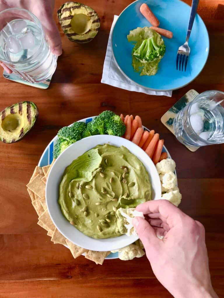 avocado hummus in a bowl surrounded by veggies and crackers with a hand dipping in a piece of cauliflower