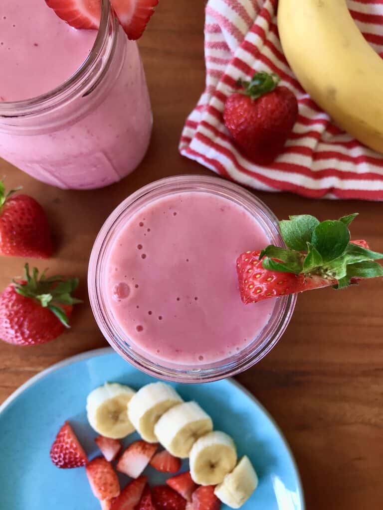 an easy smoothie recipe made with strawberries and milk in glass mason jars surrounded by strawberries, sliced banana and a red striped towel