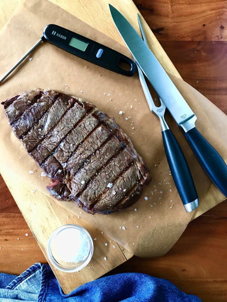 grilled steak on parchment paper on a wooden cutting board next to a bowl of salt, a carving set, thermometer and denim towel