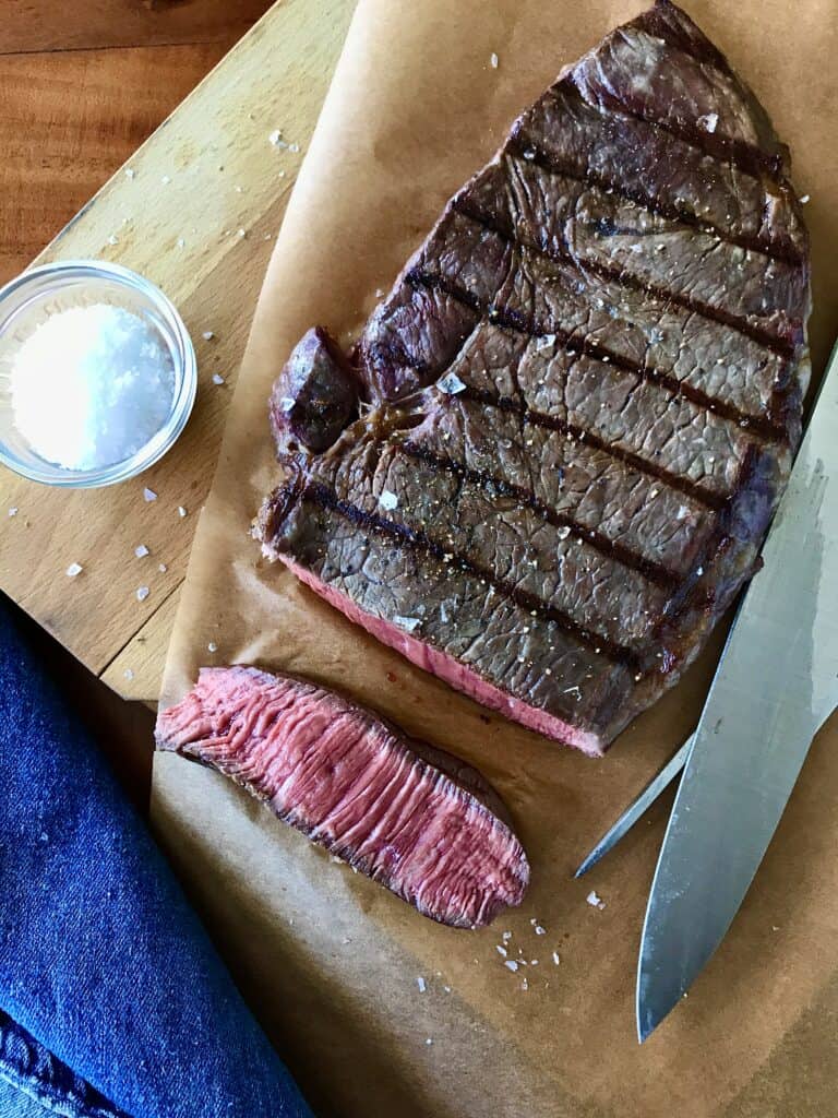 grilled steak on parchment paper on a wooden cutting board next to a bowl of salt, a carving set, thermometer and denim towel