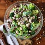 dairy free broccoli salad surrounded by raisins, sunflower seeds, a spoon and bacon