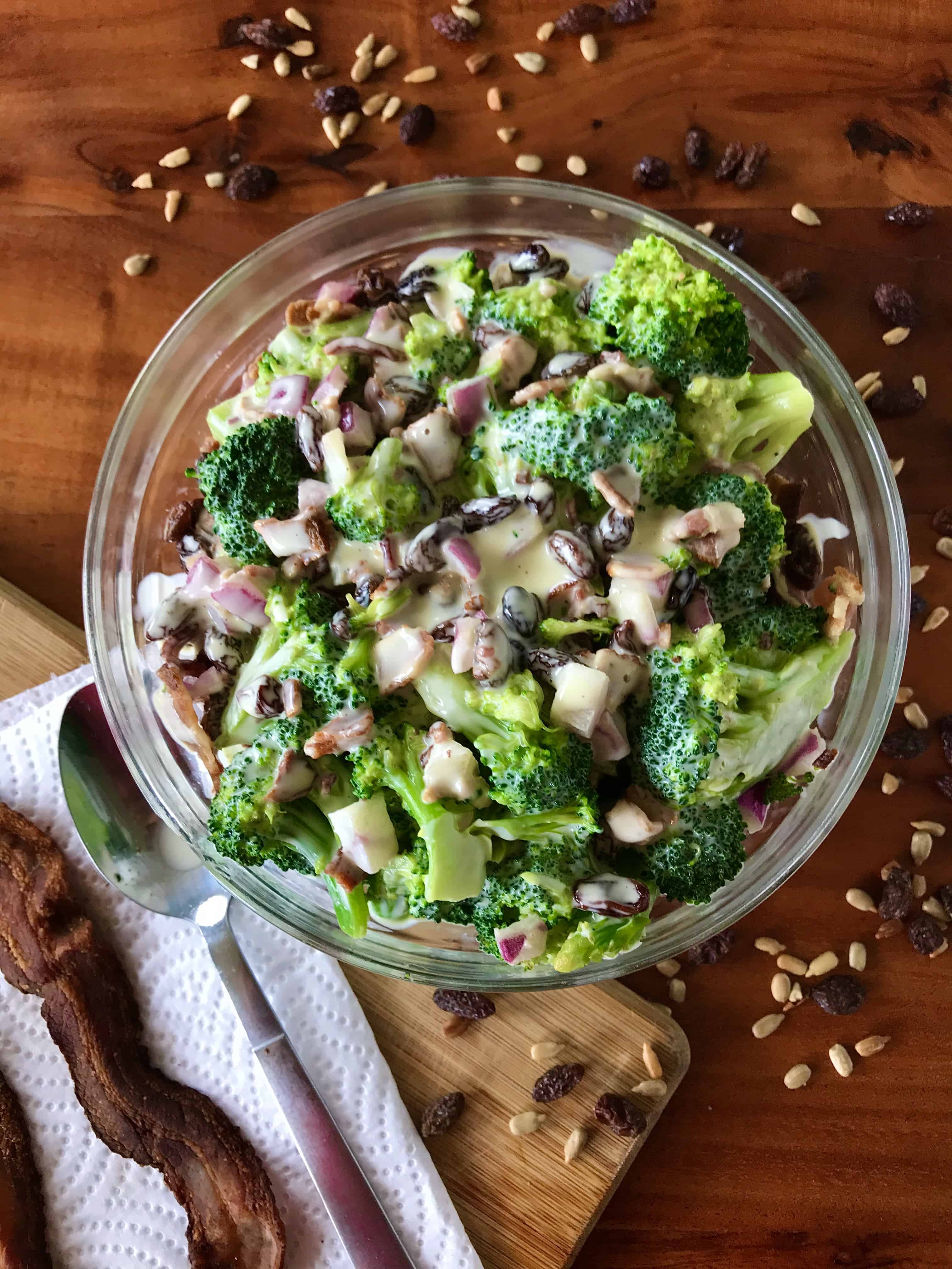 dairy free broccoli salad surrounded by raisins, sunflower seeds, a spoon and bacon