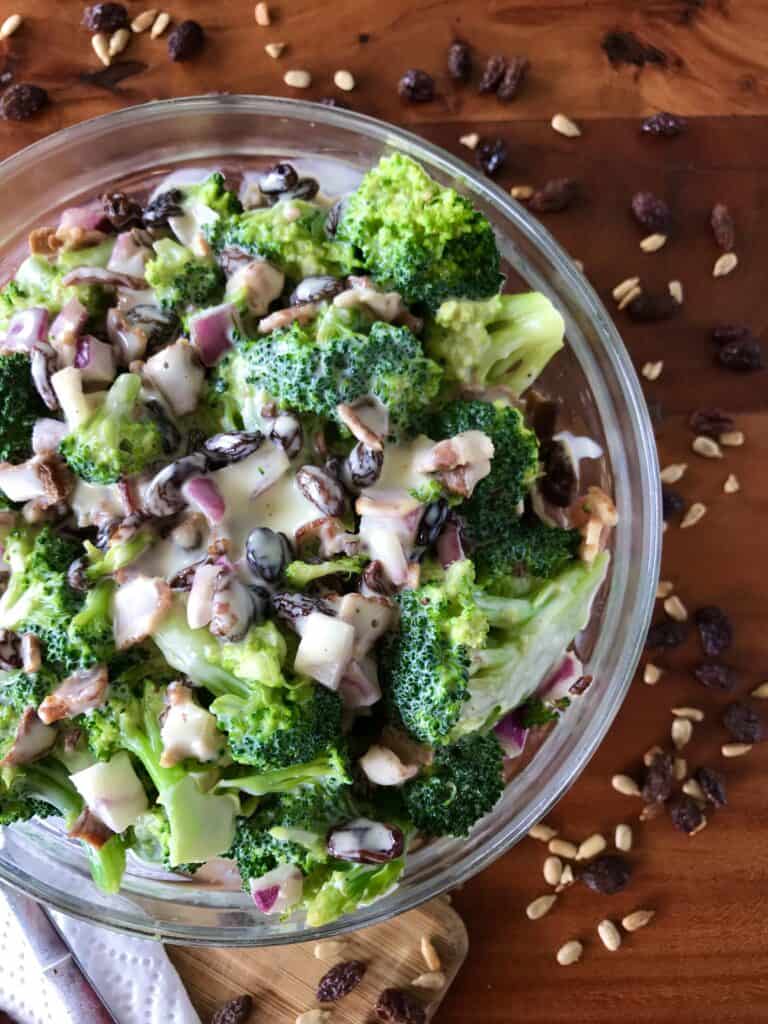 healthy broccoli salad with bacon in a glass bowl surrounded by raisins and sunflower seeds