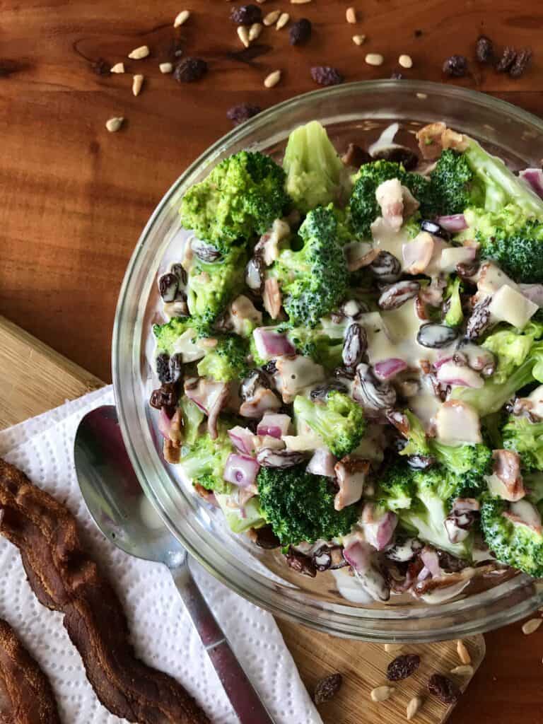 broccoli salad with bacon and raisins in a glass bowl surrounded by bacon, raisins, sunflower seeds and a spoon