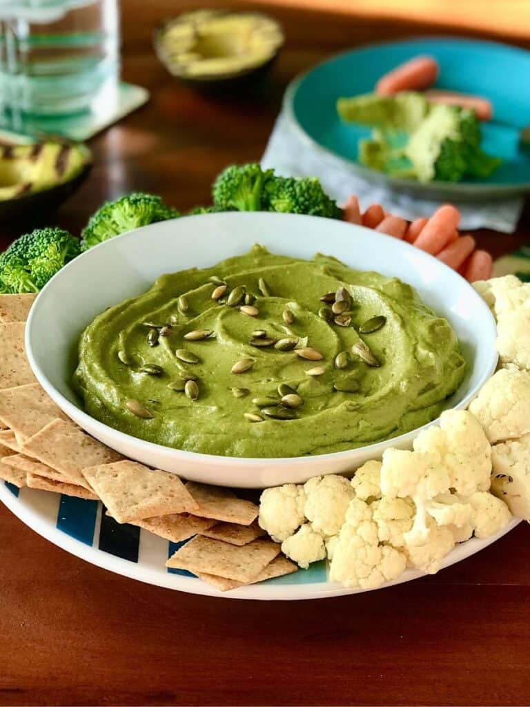 grilled avocado hummus in a bowl surrounded by veggies and crackers