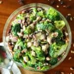 light and easy broccoli salad with bacon and raisins in a glass bowl surrounded by raisins, sunflower seeds, bacon and a spoon