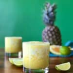 a pineapple margarita mocktail in a glass surrounded by lime and orange wedges and a pineapple