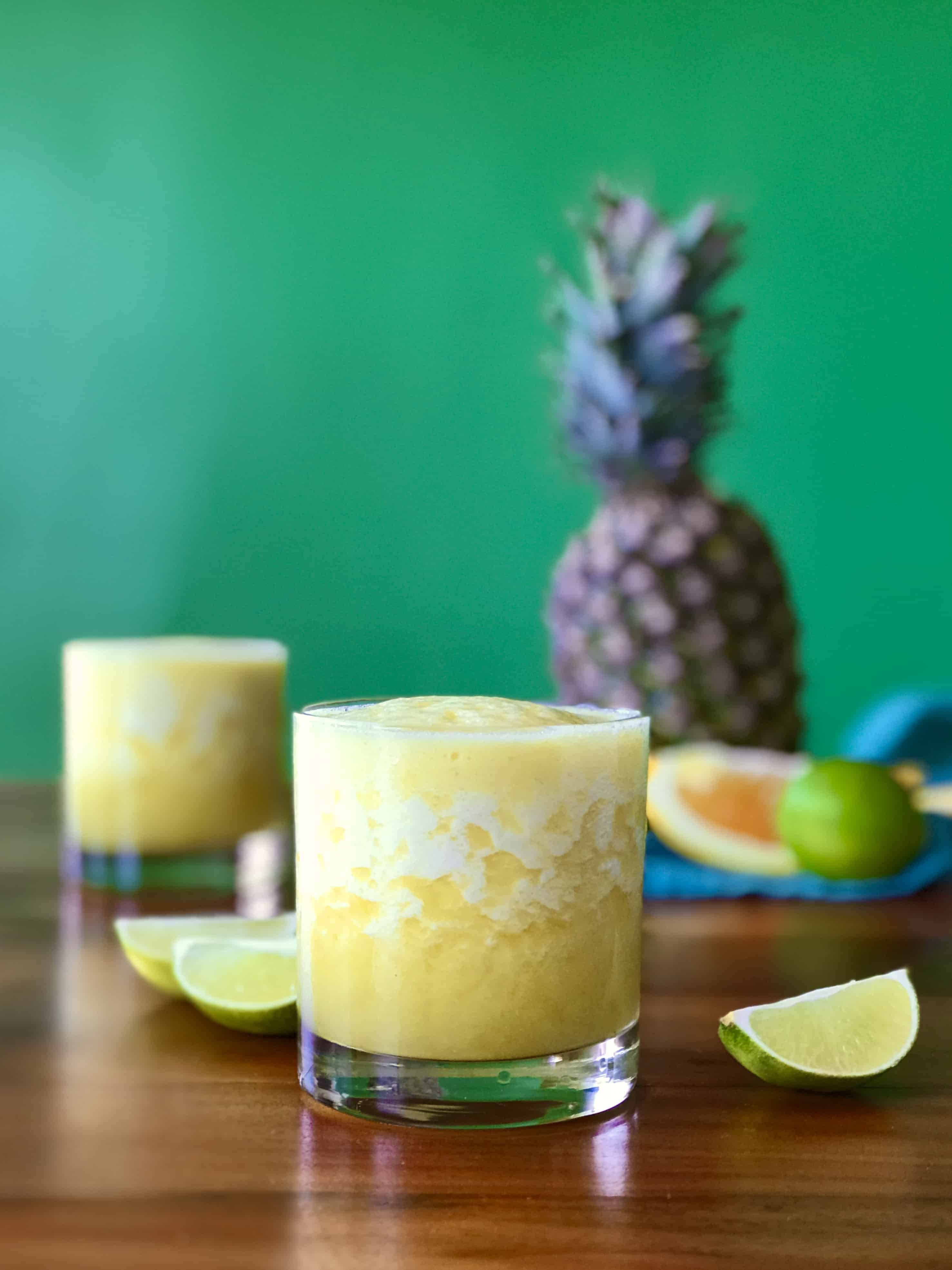 a pineapple margarita mocktail in a glass surrounded by lime and orange wedges and a pineapple