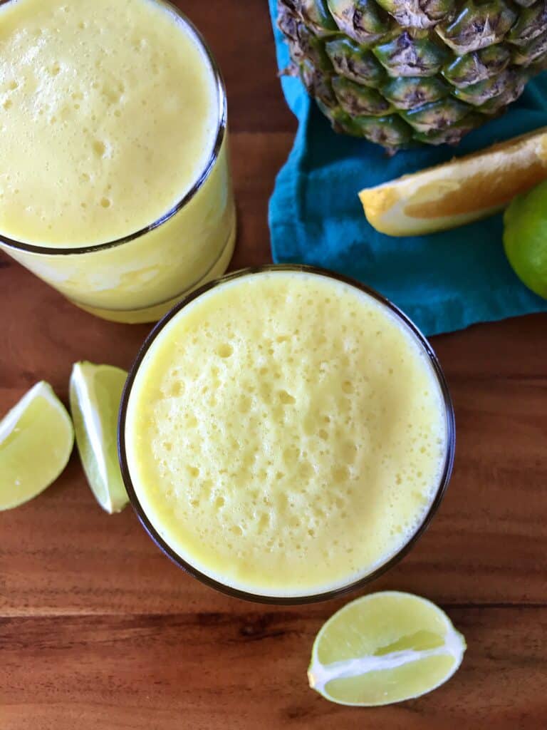 2 glasses of a frozen pineapple drink surrounded by lime and orange wedges and a pineapple