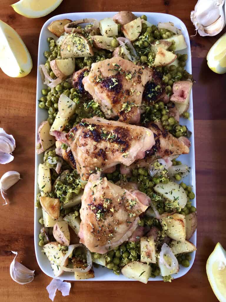 sheet pan chicken thighs with potatoes and peas on a white platter surrounded by lemon wedges and garlic cloves