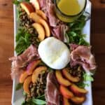 peach arugula salad with prosciutto and burrata on a white platter on a wooden table
