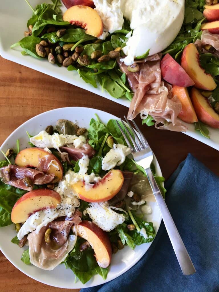 a white platter and white plate both topped with burrata, peaches, pistachios, prosciutto and arugula