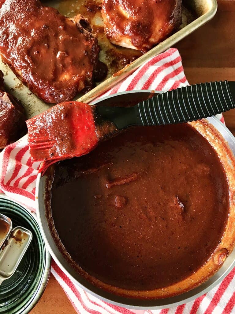 tangy bbq sauce in a pot with a brush resting on it, next to a pan of pork chops with the sauce on top