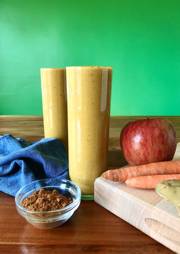 carrot and apple smoothie in a glass surrounded by carrots, apples and a bowl of cinnamon