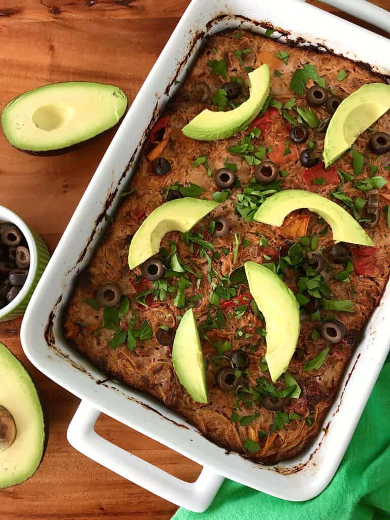 A Whole30 casserole with ground beef, peppers and more in a white baking dish, topped with avocado and sliced black olives