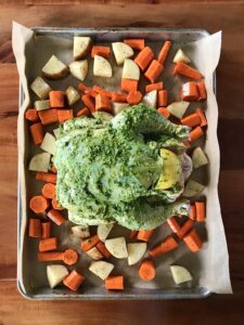 raw whole chicken coated in pesto on a baking sheet with potatoes and carrots