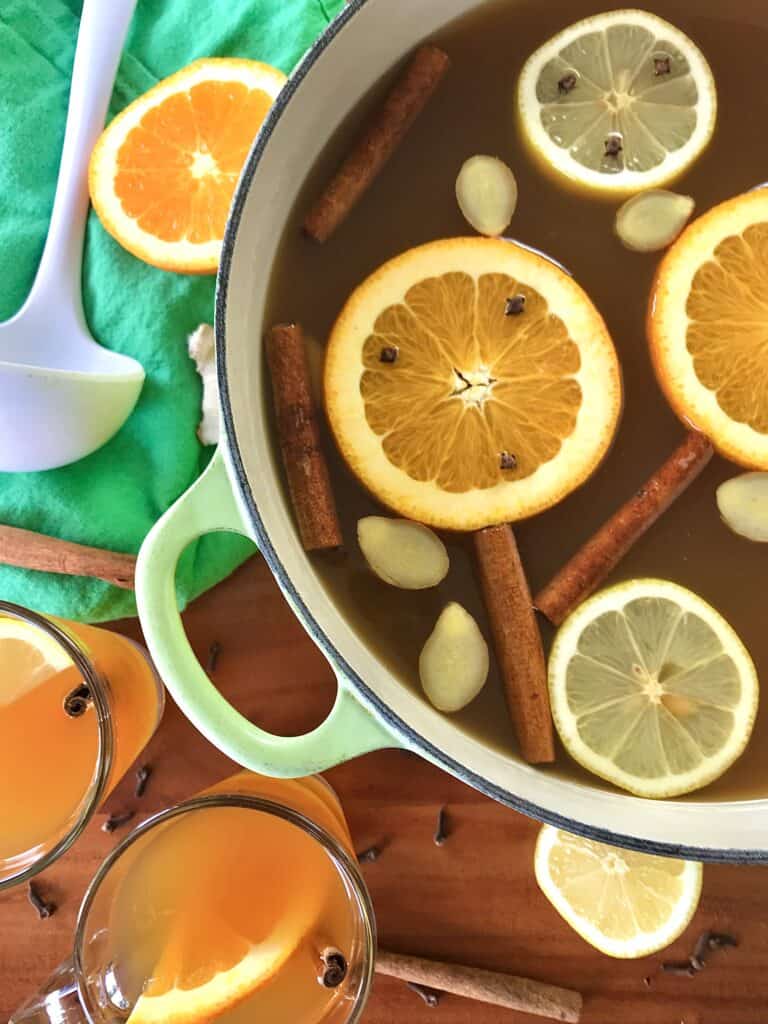 crockpot mulled cider next to glasses filled with cider, whole cloves, cinnamon sticks and a ladle