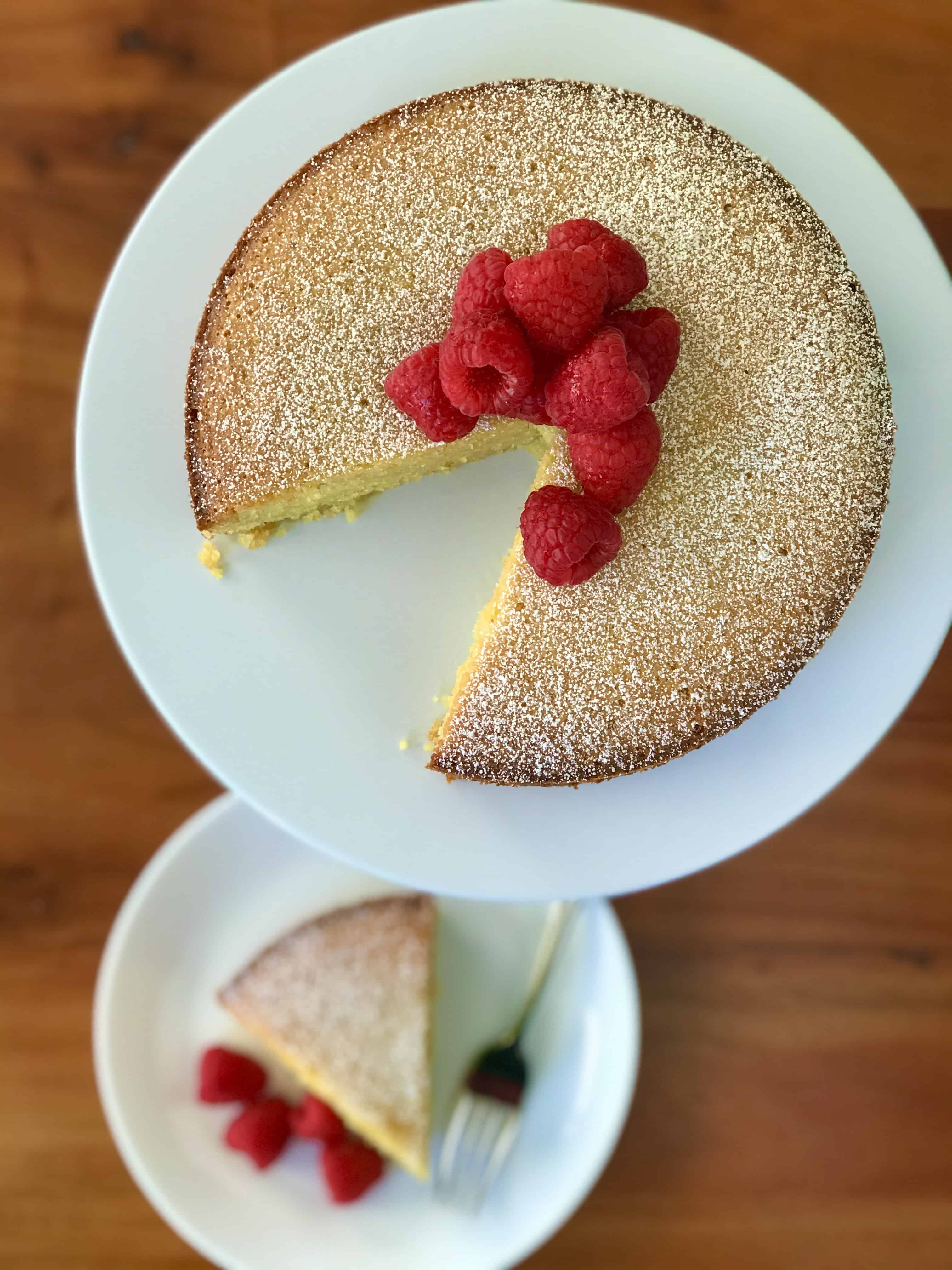 An Italian cake made with polenta and olive oil on a white platter next to a slice on a white plate with raspberries