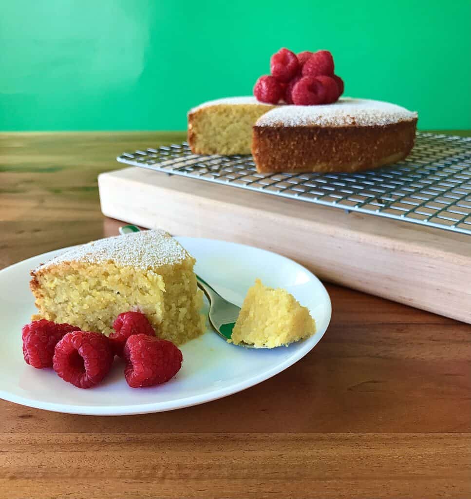 polenta cake topped with raspberries on a cutting board and a slice on a white plate on a wooden table