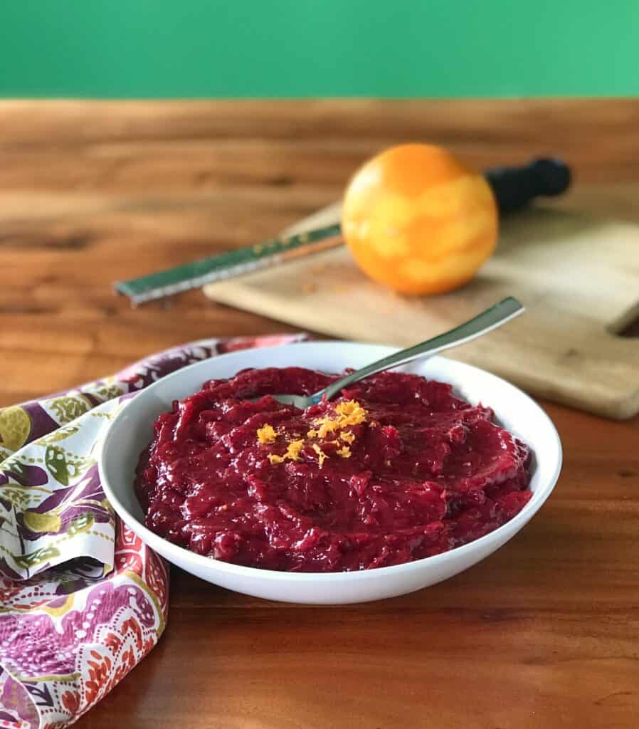 vegan cranberry sauce in a white bowl next to a napkin and a cutting board with an orange and zester