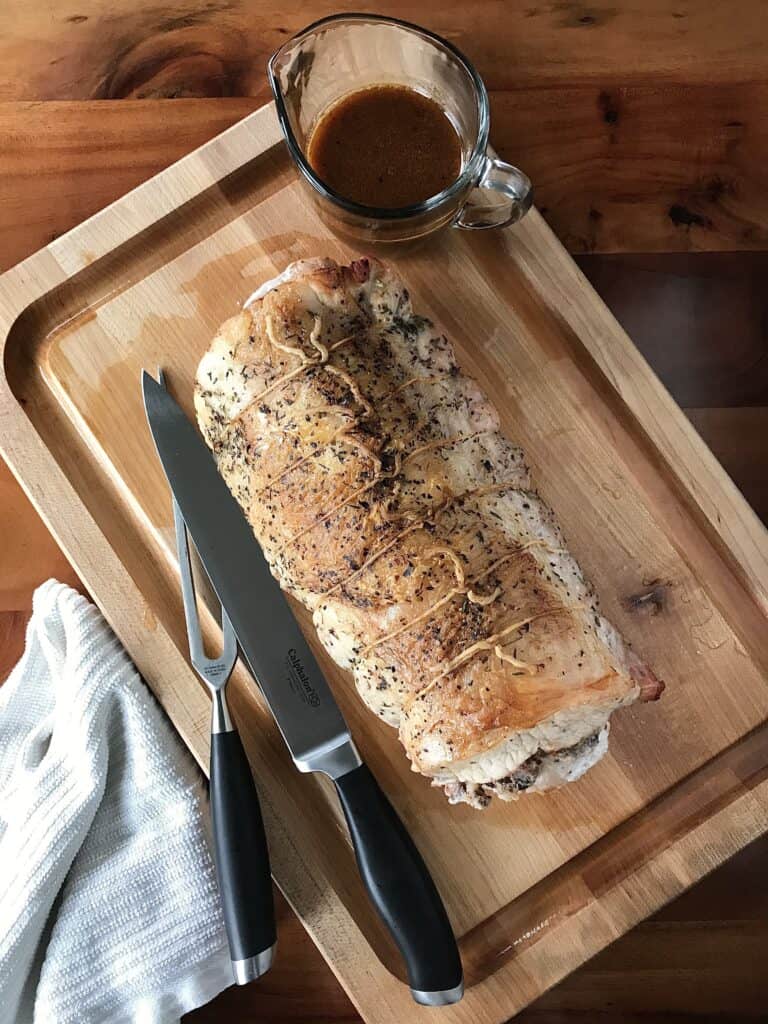 a whole roasted pork loin tied up on a cutting board next to a carving knife set