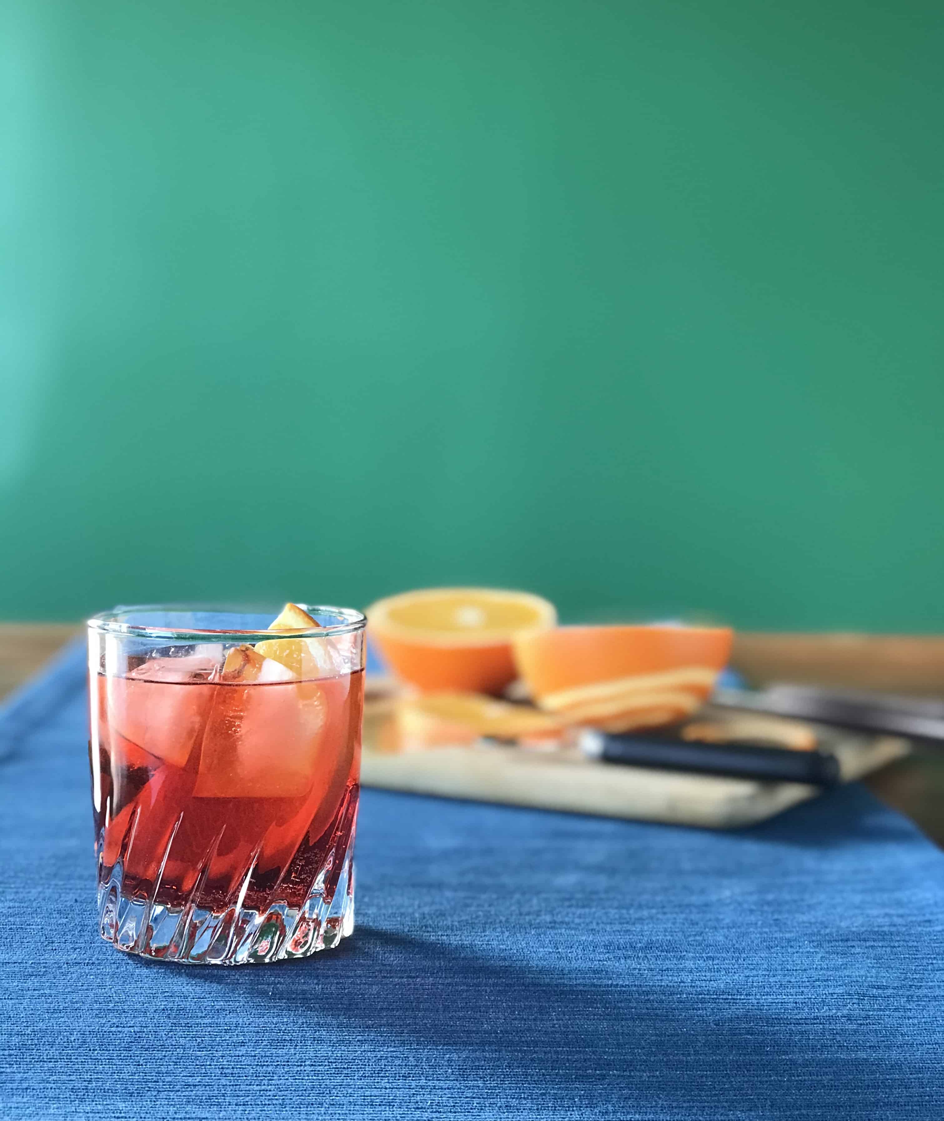 a classic Campari cocktail with vermouth and Prosecco in a glass with ice and an orange slice next to a cutting board with oranges on it