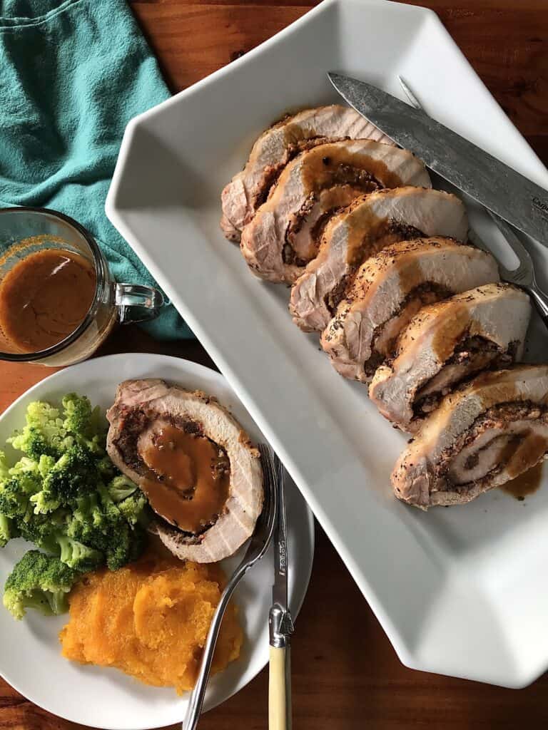 a boneless pork roast sliced on a white platter and a slice on a dinner plate covered in sauce with broccoli and mashed pumpkin