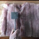 a butterflied pork loin covered in plastic wrap while a hand holding a meat mallet pounds it