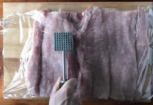 a butterflied pork loin covered in plastic wrap while a hand holding a meat mallet pounds it