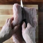 one hand using a knife to slice into a pork loin while the other hand is opening it up like a book