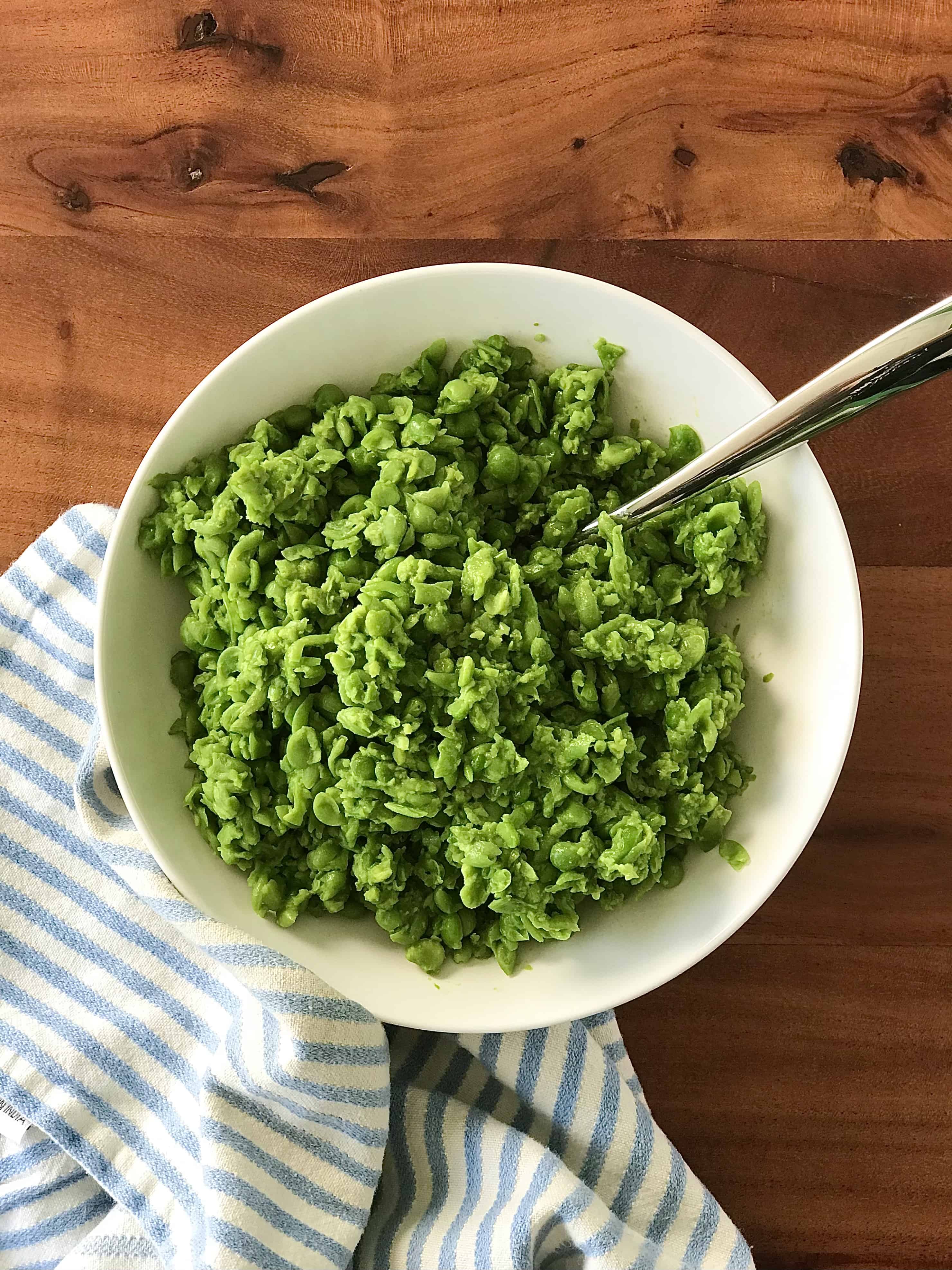 mushy peas in a white bowl on a wooden table next to a blue striped towel