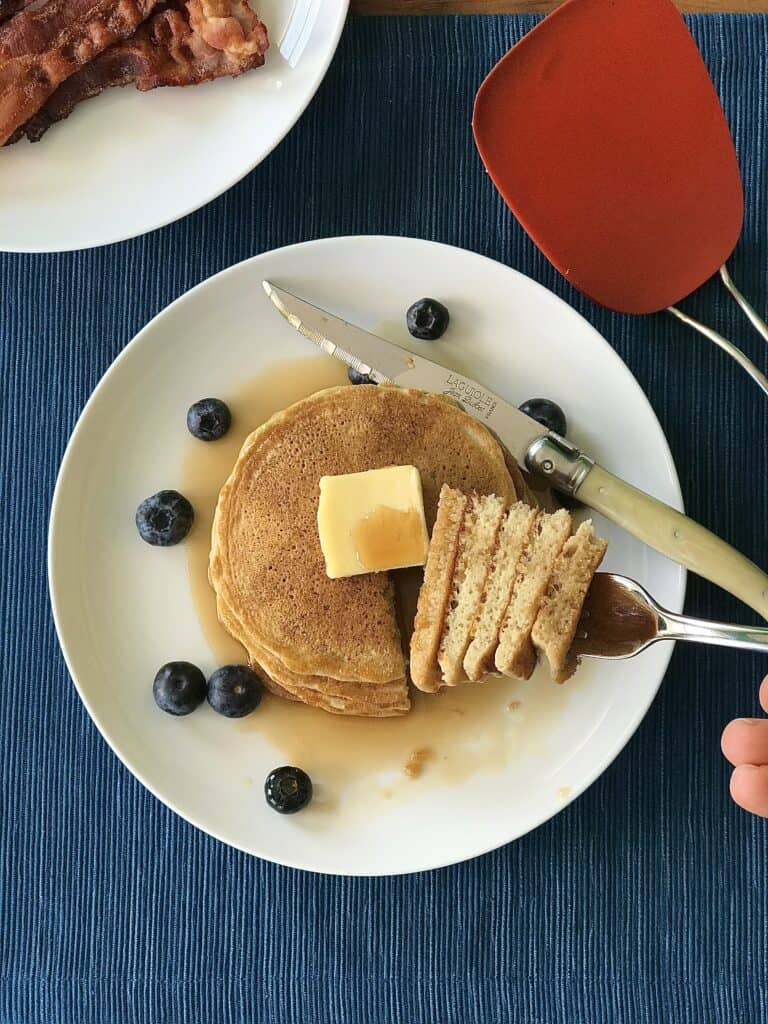 an overhead shot of paleo pancakes stacked on a white plate with blueberries, butter and maple syrup with a fork holding 5 slices