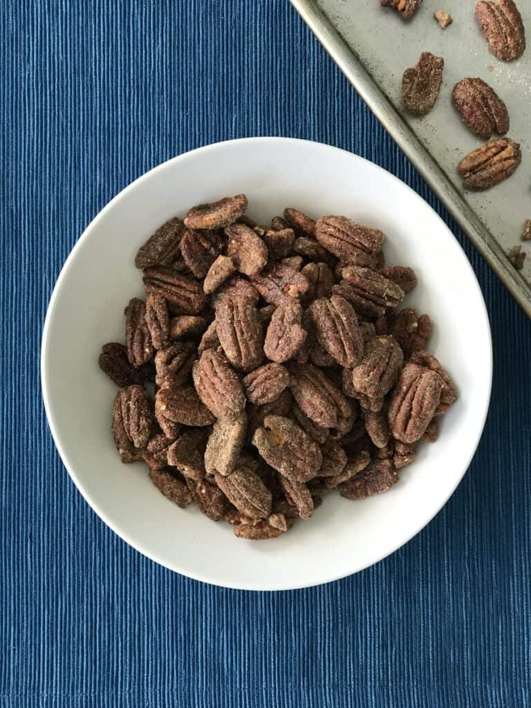 easy, healthy roasted pecans in a white bowl on a blue table runner next to a baking sheet with pecan crumbs on it