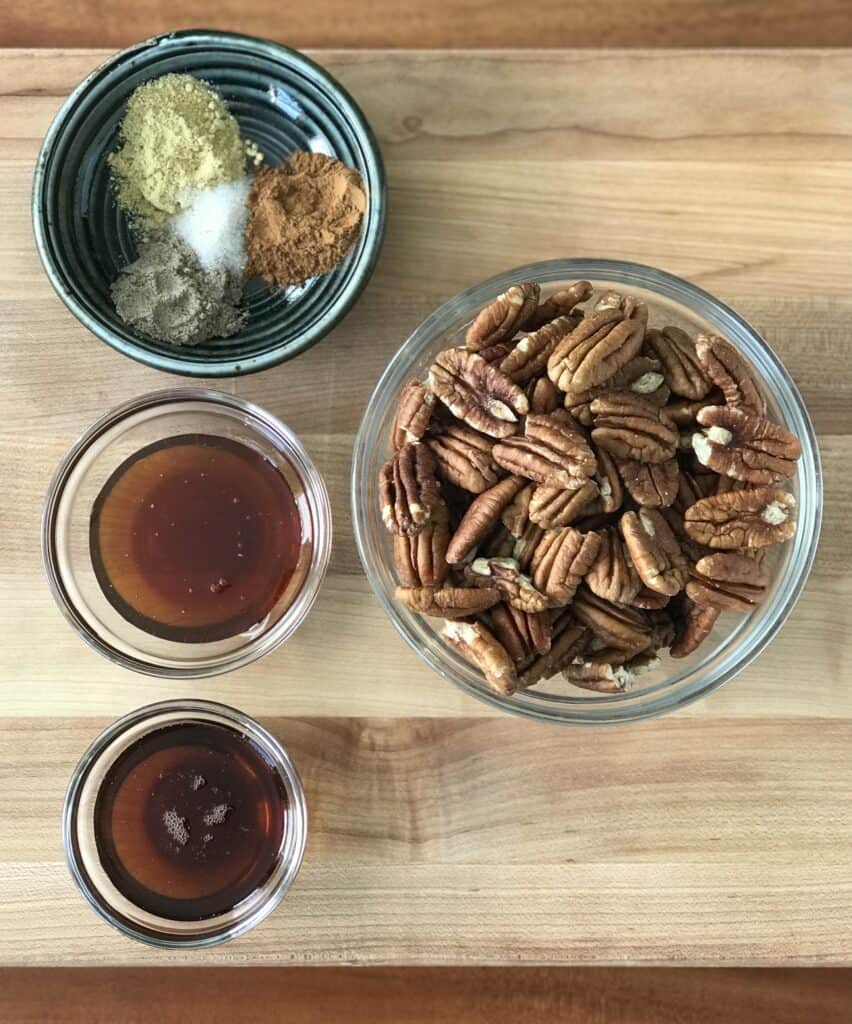 the individual ingredients to make spiced pecans in bowls on a wooden cutting board