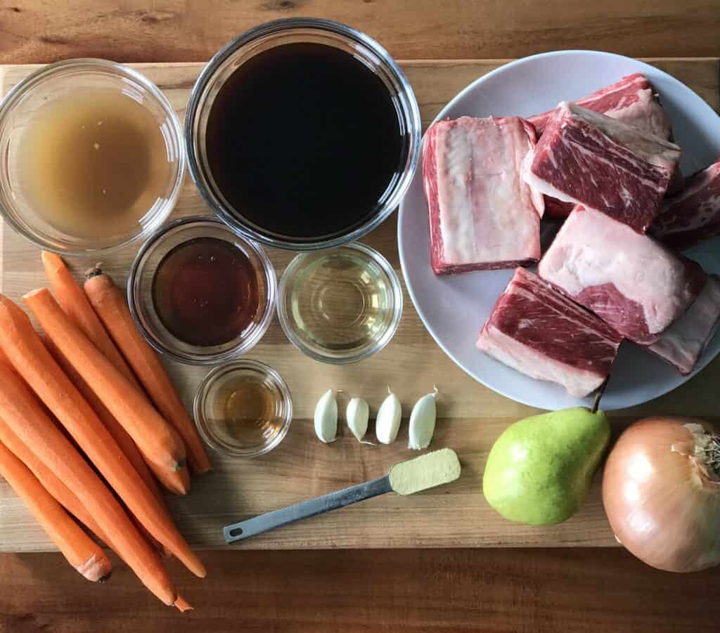 a wooden cutting board full of short ribs, carrots, an onion, a pear, condiments, garlic and a measuring spoon full of ground ginger