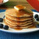 gluten-free pancakes made with coconut flour stacked on a plate with butter, maple syrup and blueberries on a white plate on a blue table runner