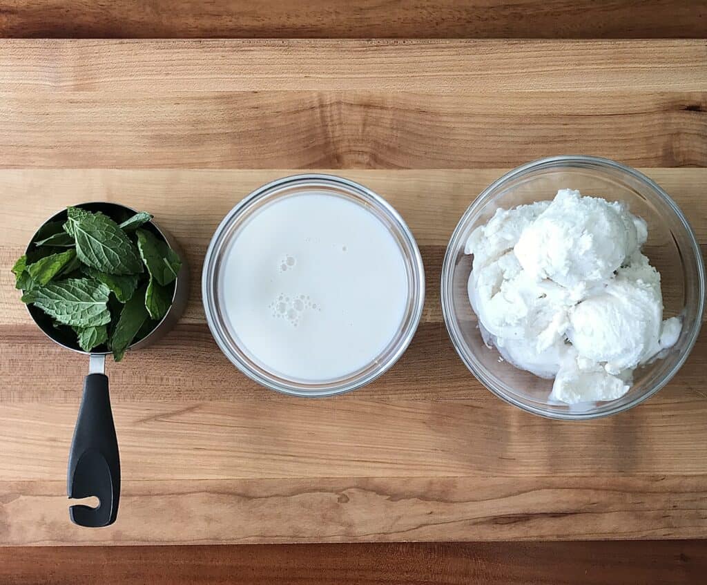 fresh mint in a measuring cup, almond milk and vanilla ice cream in glass bowls, all on a wooden cutting board