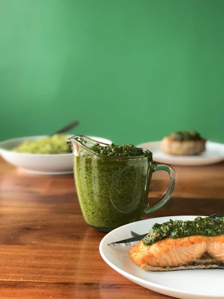 Italian pesto in a small glass pitcher next to a white plate with pesto-covered salmon on it, plus a white bowl with pesto spaghetti and a white plate with a pesto-covered turkey burger, all on a wooden table