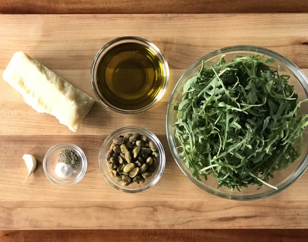 arugula, olive oil, salt and pepper, and pistachios in glass bowls next to a block of Parmesan cheese and a garlic clove, all on a wooden cutting board on a wooden table