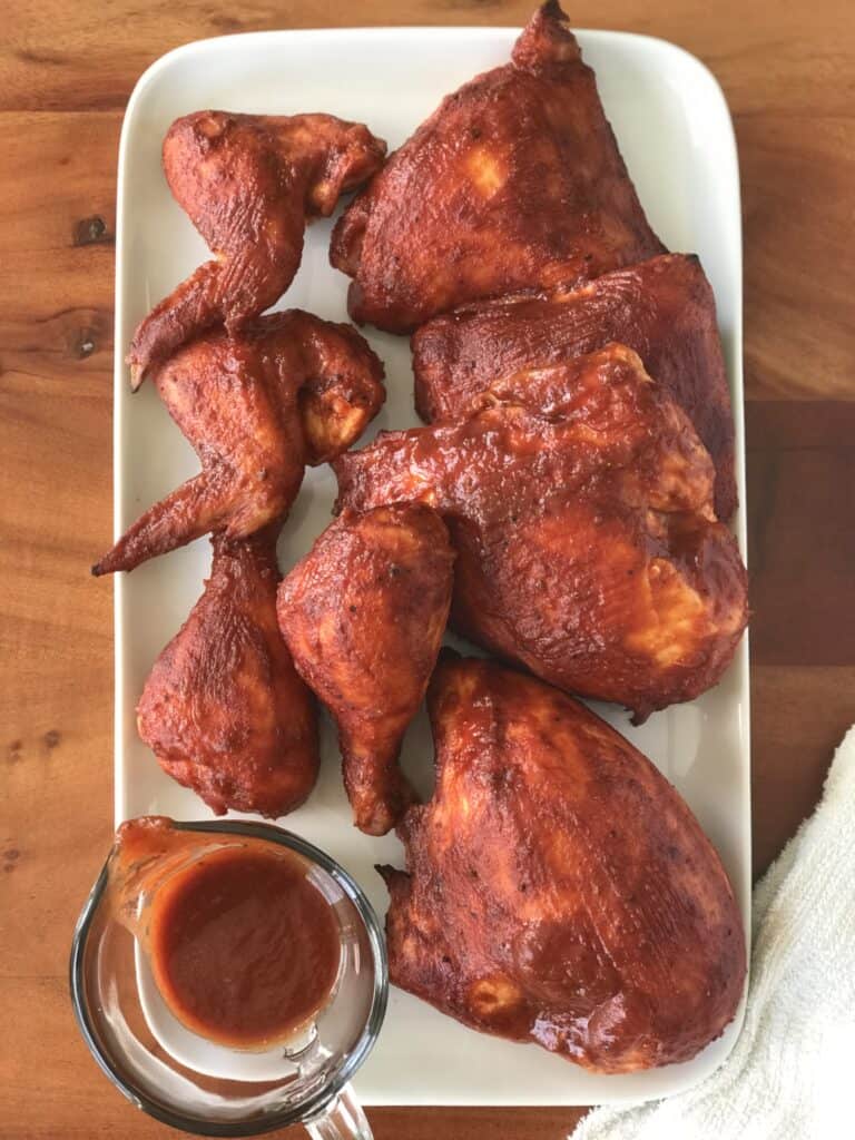 Chicken parts brushed with BBQ sauce and a small glass pitcher with more sauce sit on a white serving platter next to a white towel on a wooden table