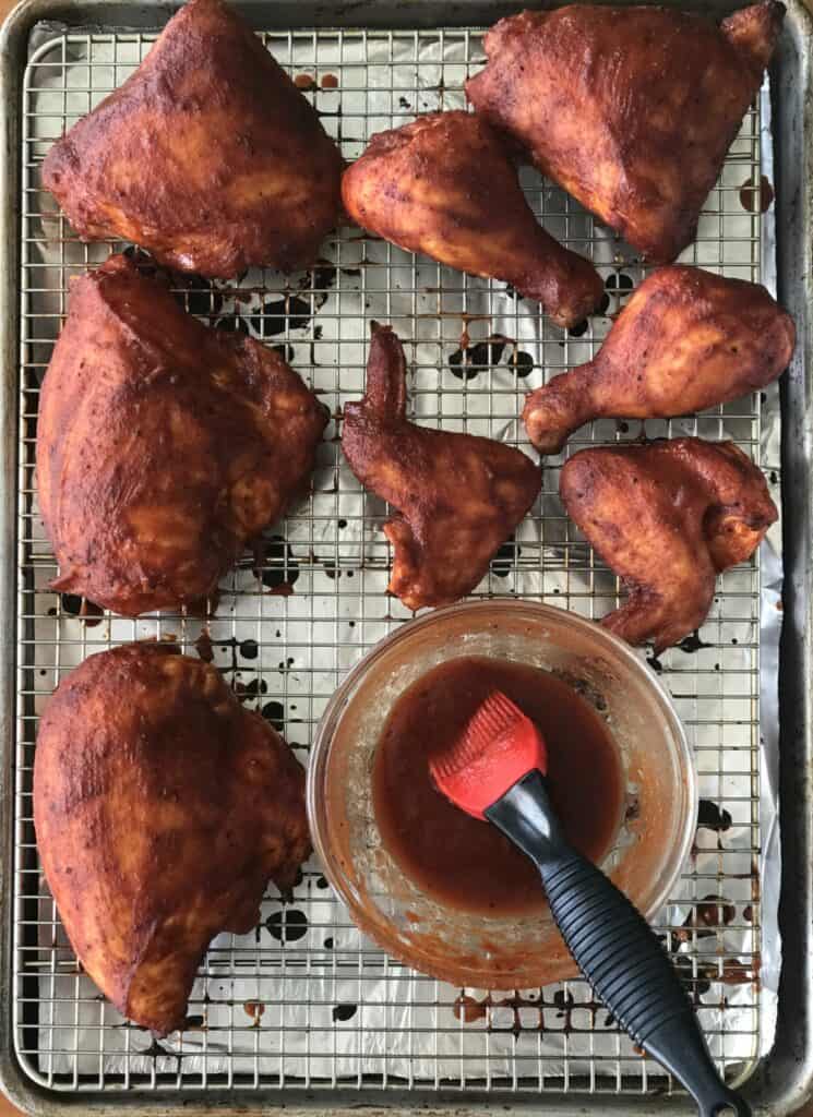 Chicken parts brushed with BBQ sauce and a glass bowl with more sauce and a silicone brush in it sit on a rack on top of a foil-lined baking sheet