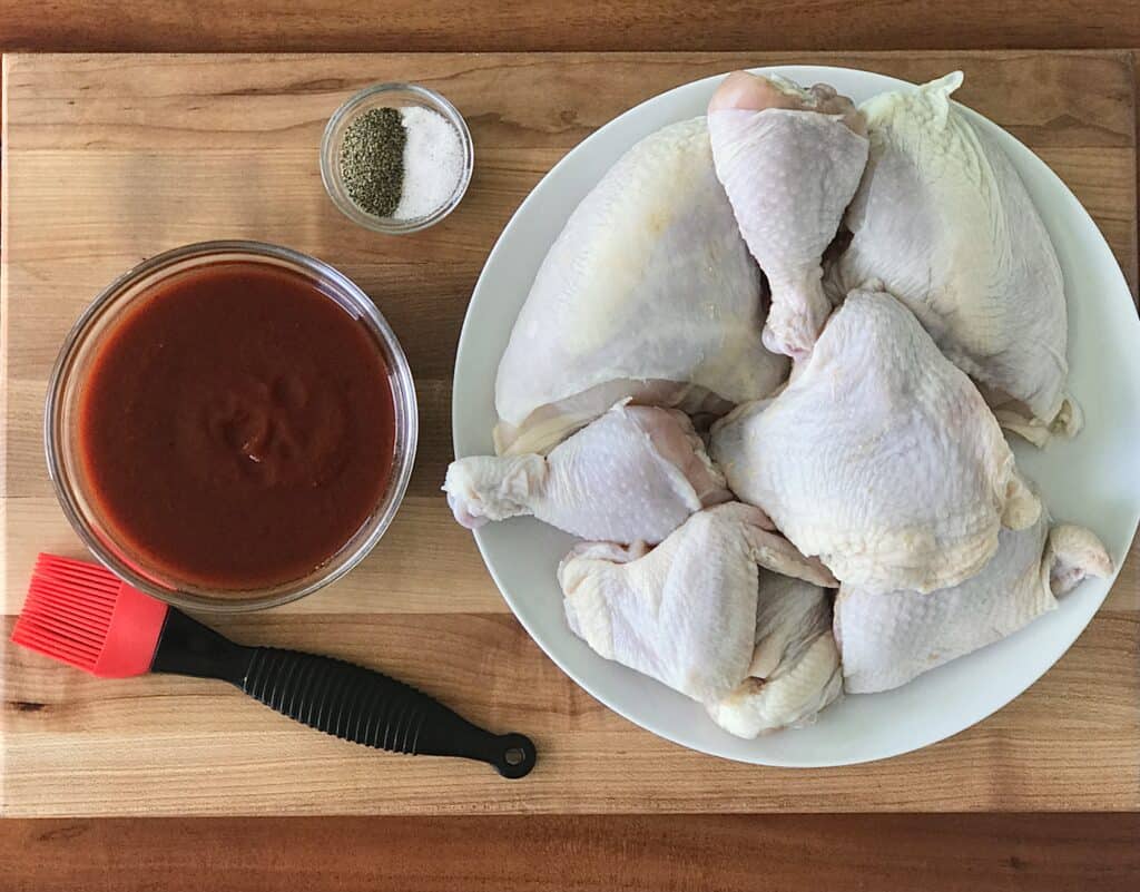 a white plate with a whole cut-up chicken, a glass bowl with BBQ sauce, a small bowl with salt and pepper, and a silicone basting brush are all sitting on a wooden cutting board