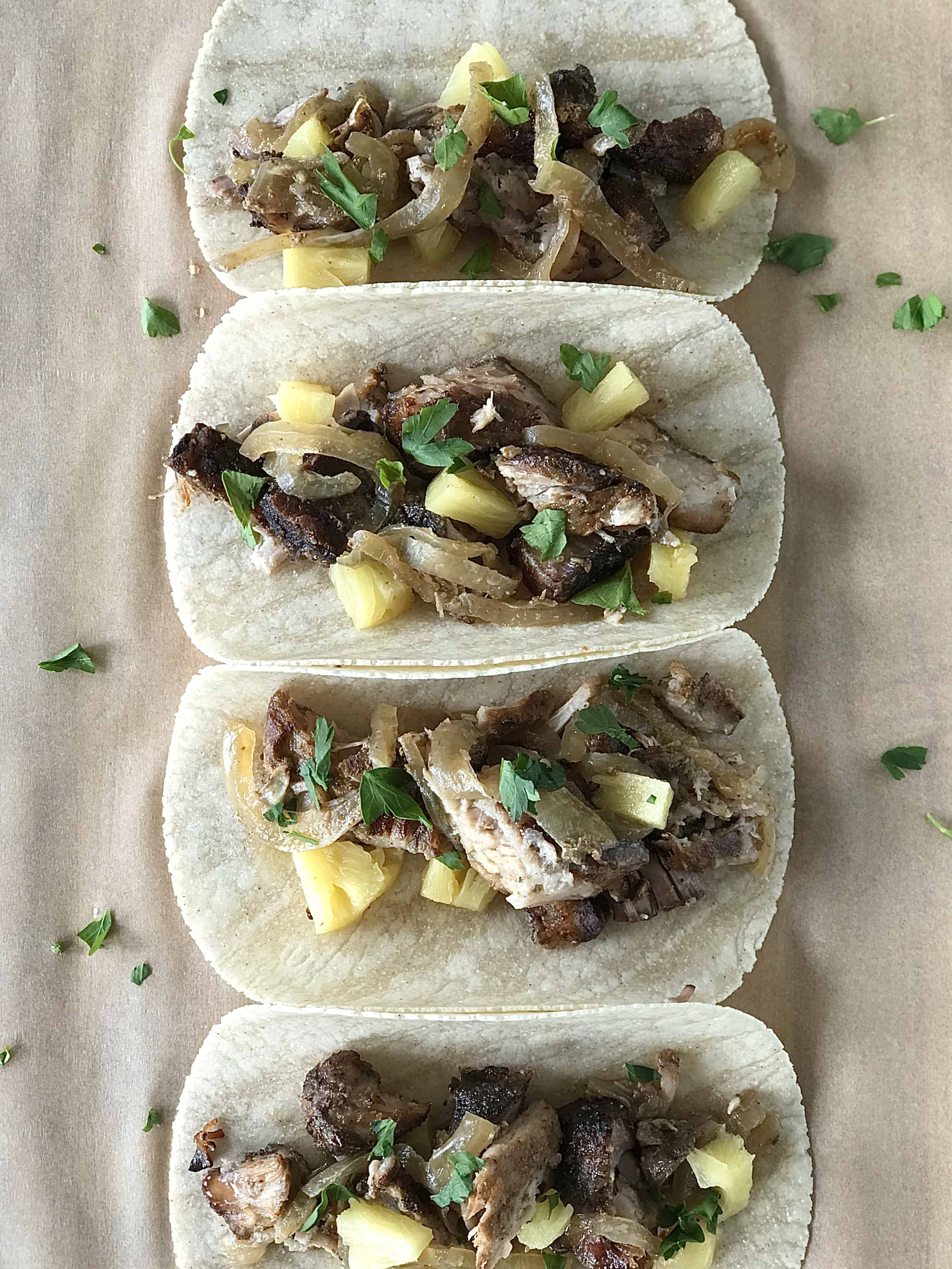 a closeup of Mexican pork tacos with onions, pineapple and cilantro in tortillas lined up on parchment paper